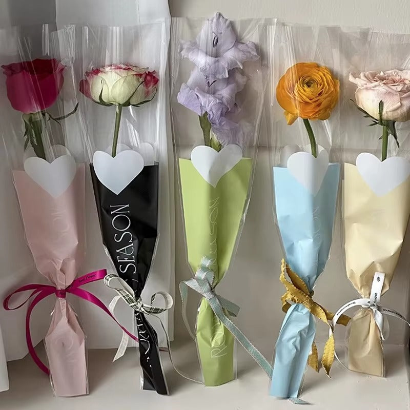 Five wrapped single flowers arranged side by side in clear plastic sleeves with paper cone wraps in pink, black, light green, light blue, and cream, each tied with a ribbon and topped with a white heart-shaped card.